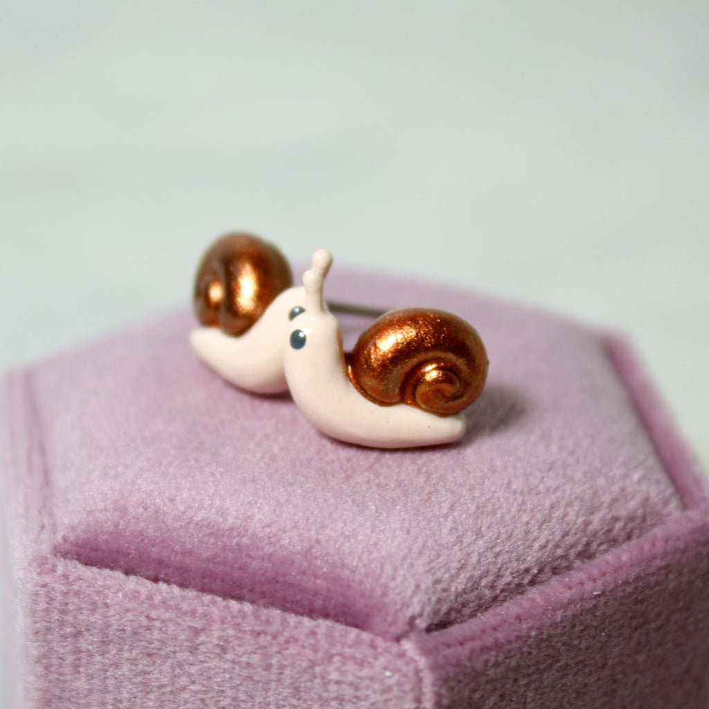 A pair of snail earrings with bronze shells sit on top of a purple jewelry box