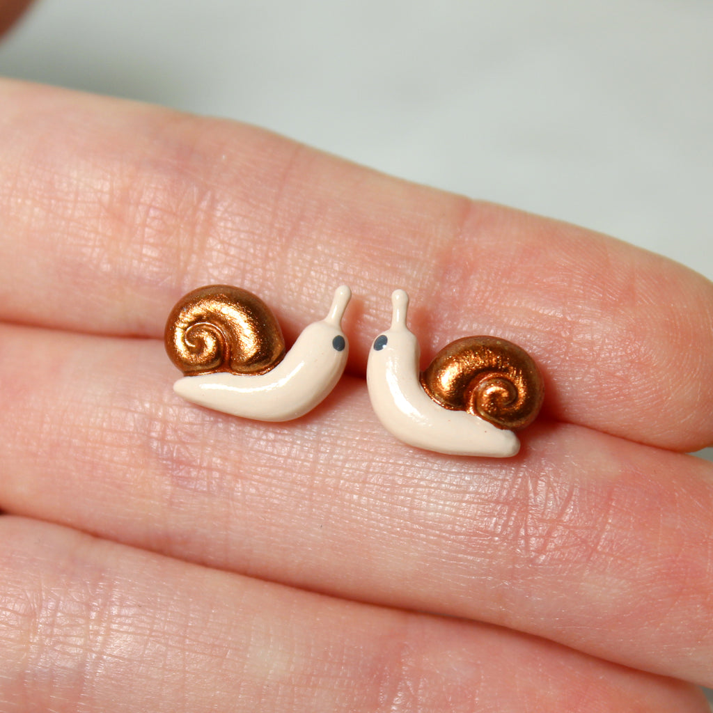 A hand holds a pair of snail stud earrings with sparkly bronze shells