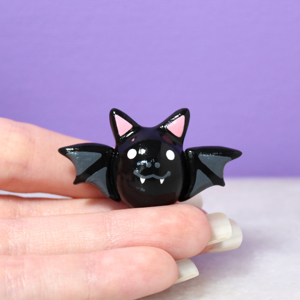 A hand holding a black bat figurine. The bat has a big, toothy smile and outstretched wings. It is about one inch tall.