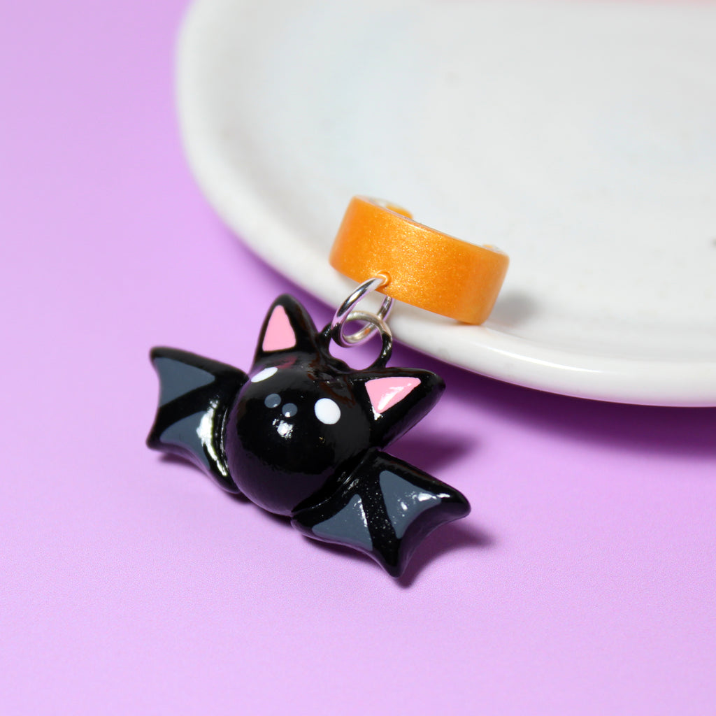 A black bat charm hangs from a sparkly orange ear cuff. The ear cuff rests against a white jewelry dish.