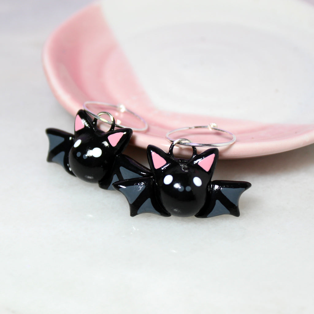 A pair of bat hoop earrings rest against a pink and white jewelry dish.