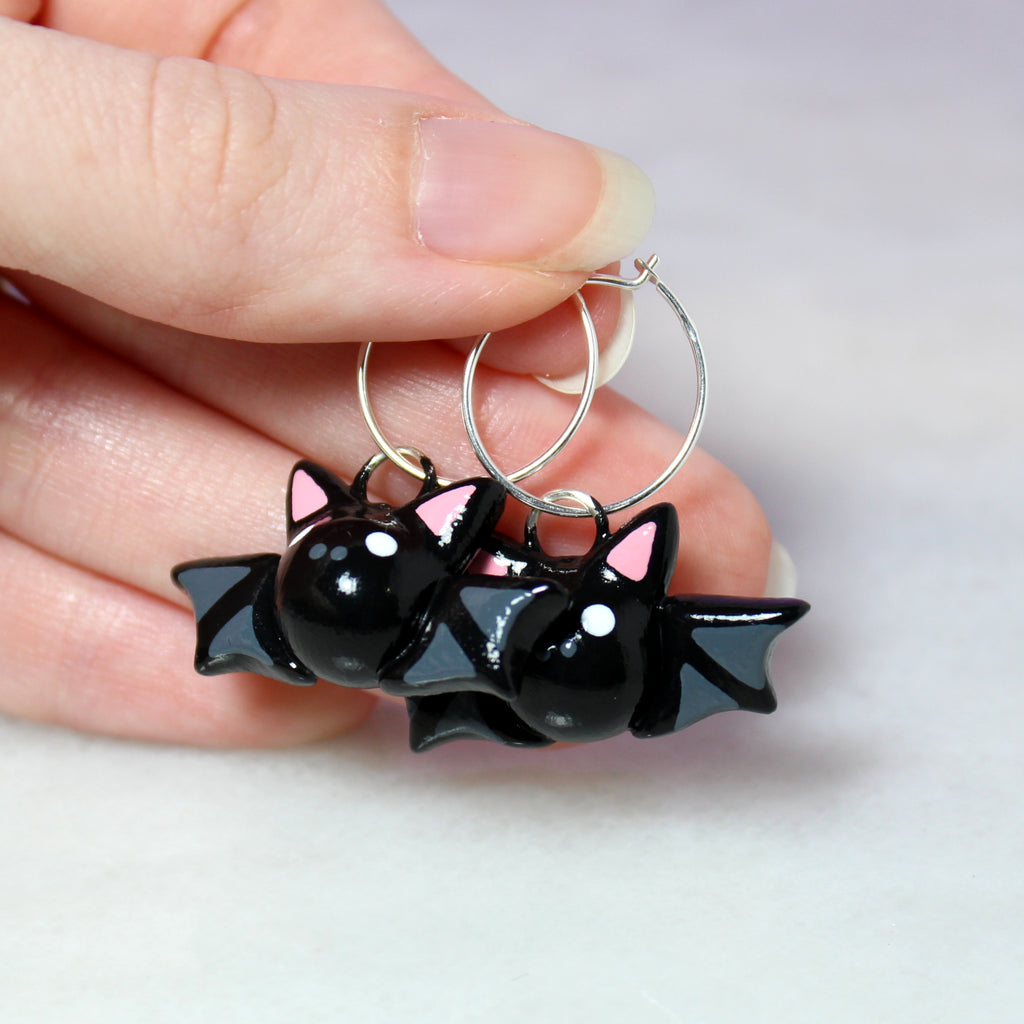 A hand holding a pair of bat hoop earrings. The bat charms are about 3/4 of an inch tall, and the hoops are 19mm wide.