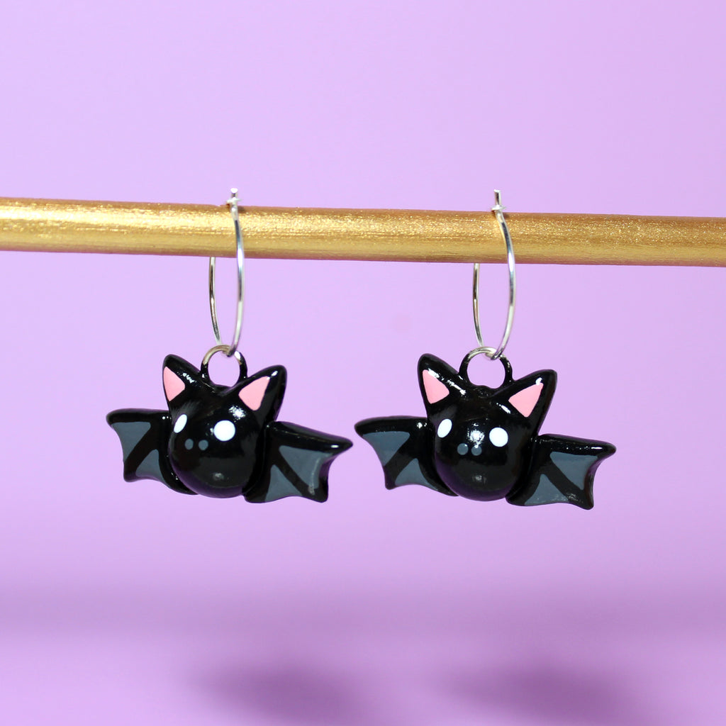 A pair of bat hoop earrings hanging from a gold bar. The background is purple.