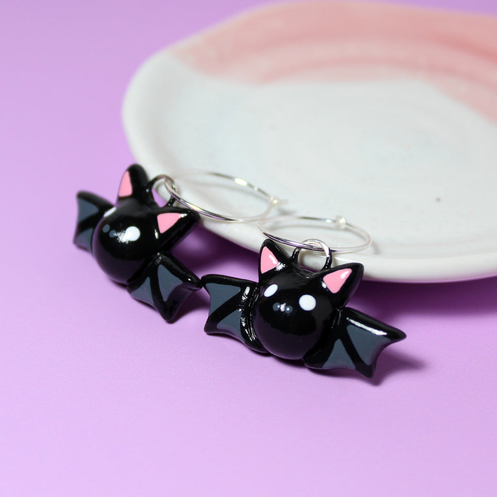 A pair of bat hoop earrings rest against a pink and white jewelry dish. The background is purple.