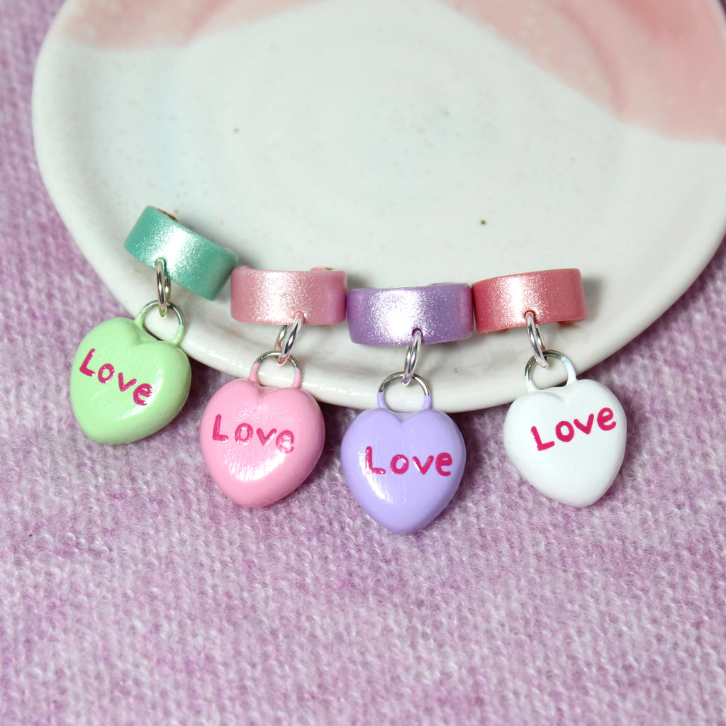 Four candy heart ear cuffs are lined up against a jewelry dish. Each candy heart charm has the word "Love" written on them.