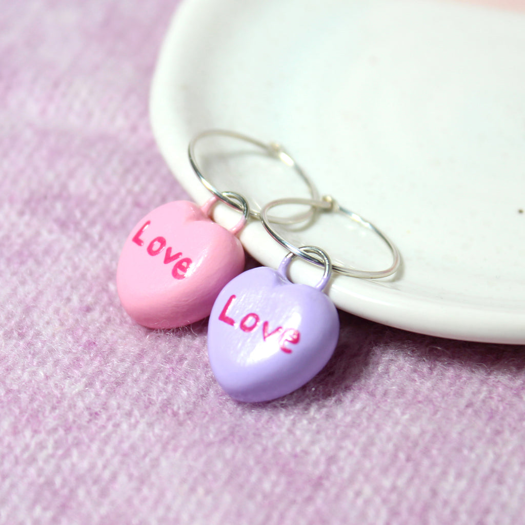 A pair of mismatched candy heart hoop earrings rest against a white jewelry dish.
