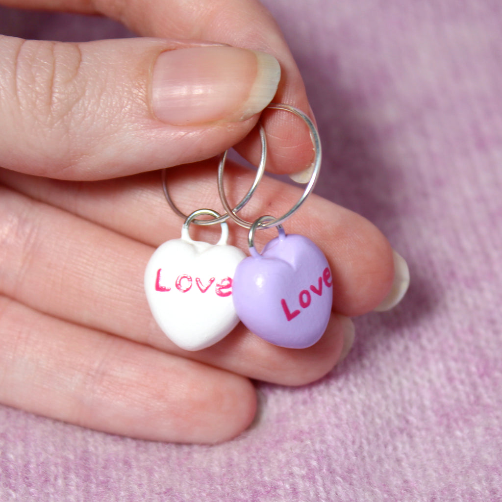 A hand holds a pair of candy heart hoop earrings. The sterling silver hoops are 19mm wide and the candy heart charms are about 3/4 of an inch tall.