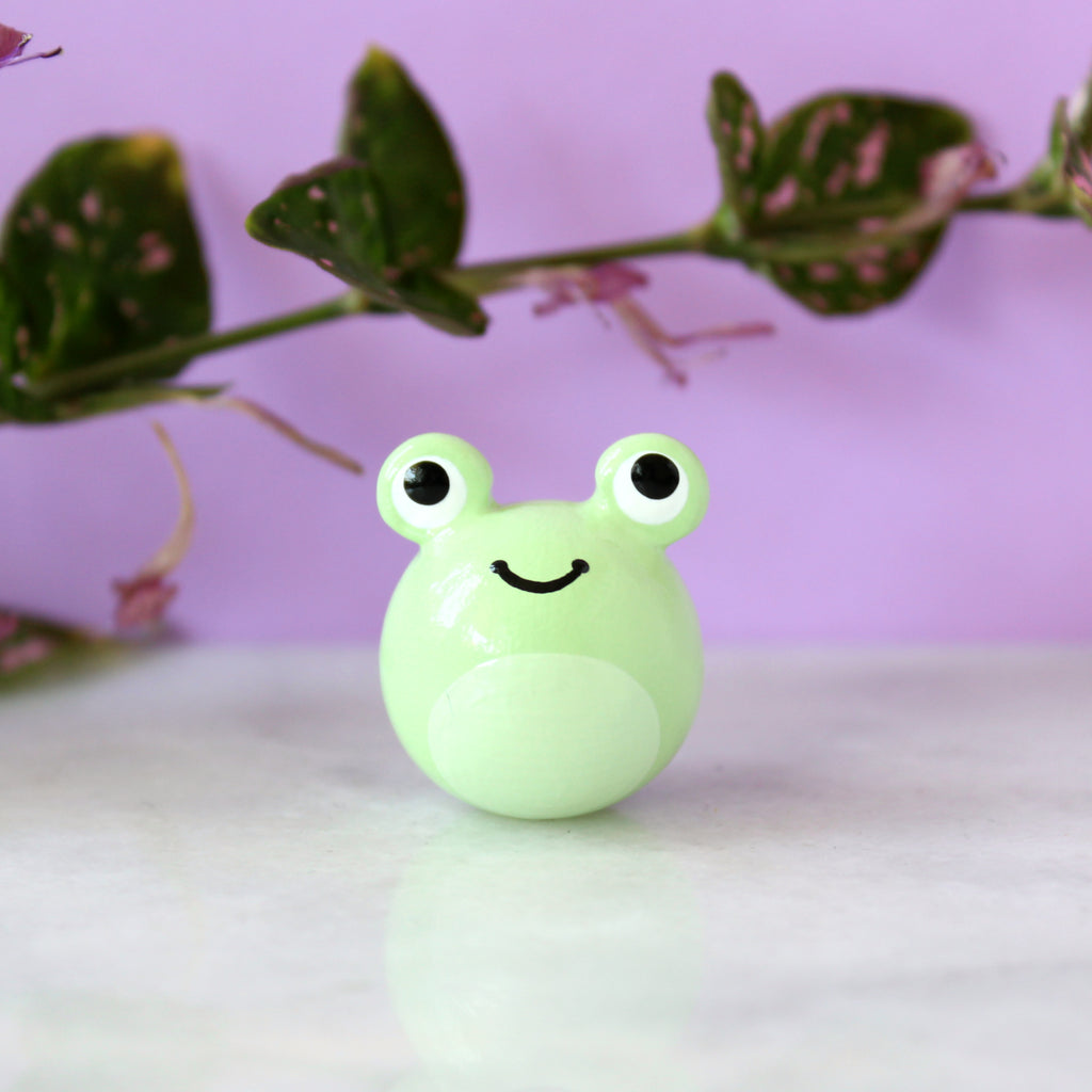 A chubby green frog figurine with big eyes and a wide happy smile. Behind the figurine is a purple background and green leaves.
