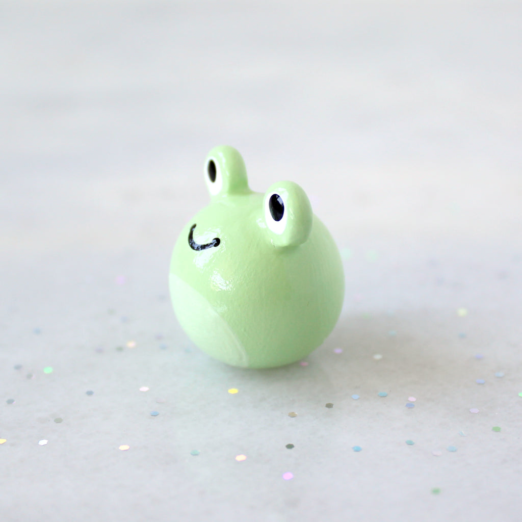 Side view of a chubby green frog figurine with big eyes and a wide happy smile. The figurine is sitting on a marble surface with rainbow glitter.