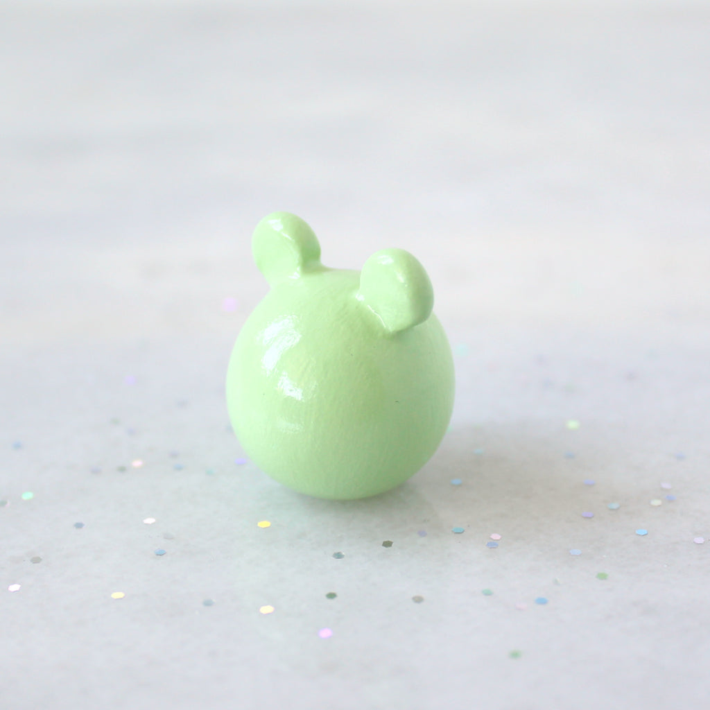 Back view of a chubby green frog figurine. The figurine is sitting on a marble surface with rainbow glitter.