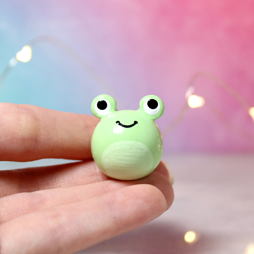 A hand holding a chubby green frog figurine. The frog has big eyes and a wide happy smile. It is about 1 inch tall. The background is blue and red with fairy lights.