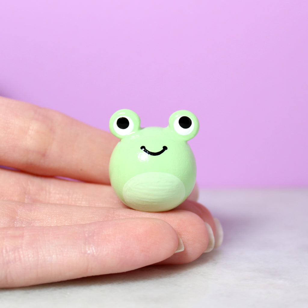 A hand holding a chubby green frog figurine. The frog has big eyes and a wide happy smile. It is about 1 inch tall. The background is purple.