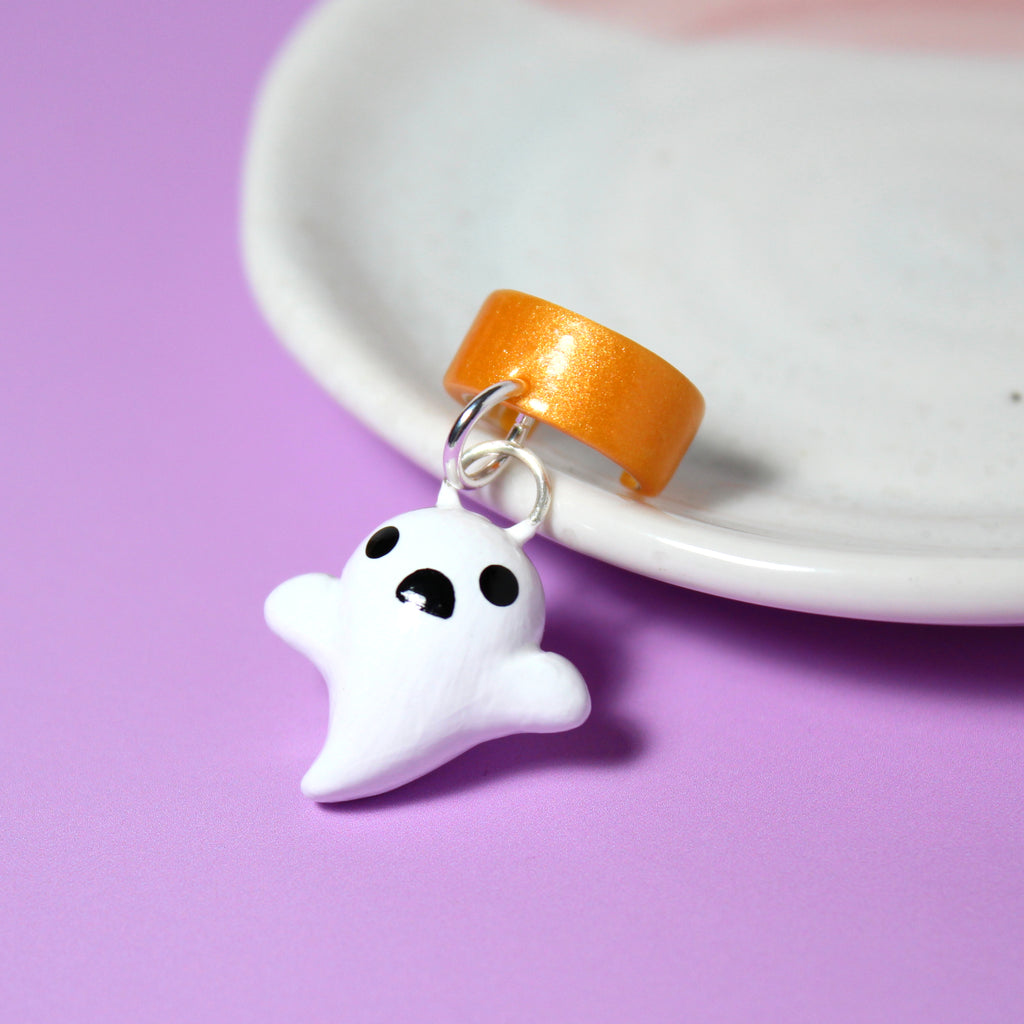 A ghost charm hangs from a sparkly orange ear cuff. The ear cuff rests against a white jewelry dish.