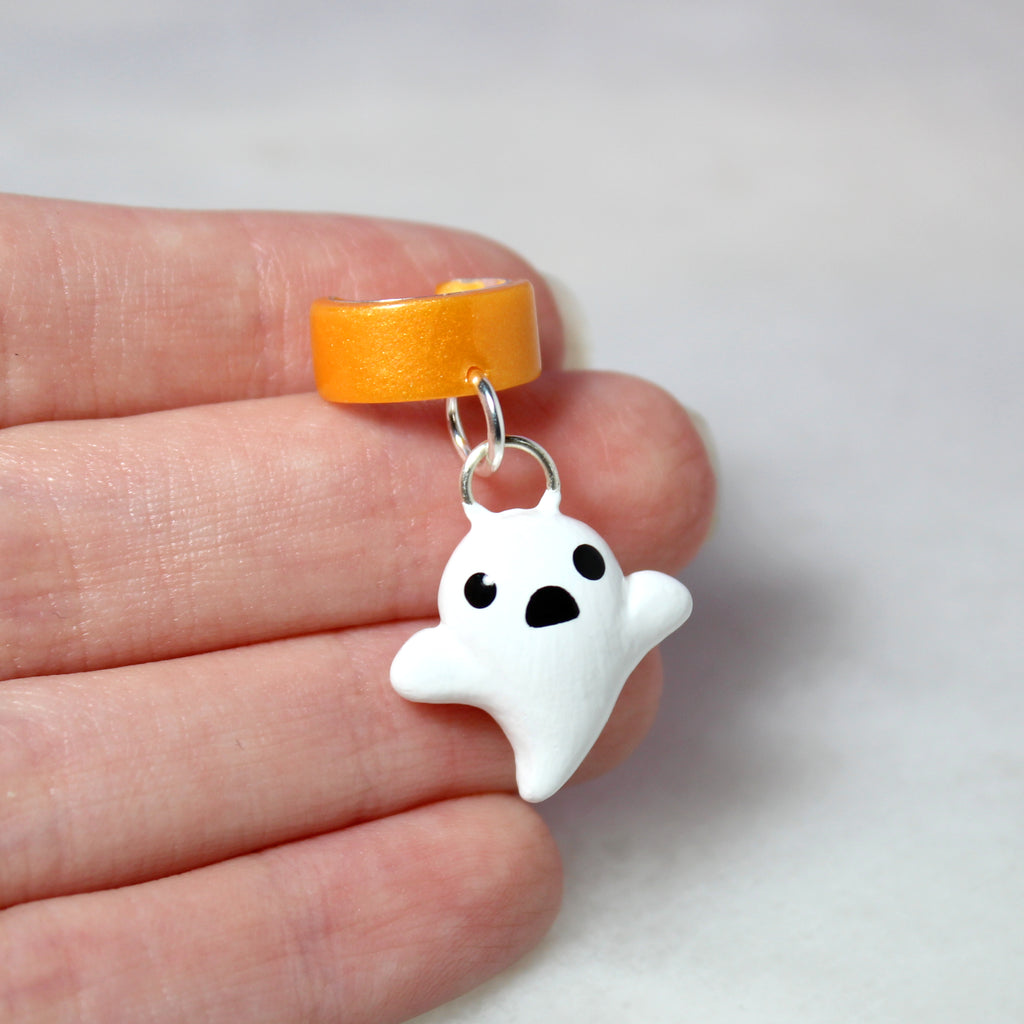 A hand holding a ghost ear cuff. The ghost charm is about 3/4 of an inch tall.