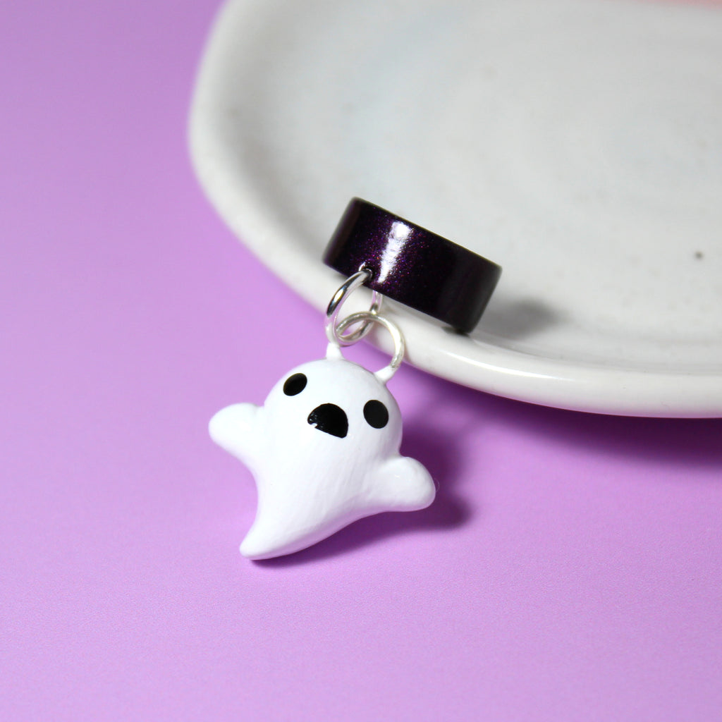 A ghost charm hangs from a sparkly dark purple ear cuff. The ear cuff rests against a white jewelry dish.