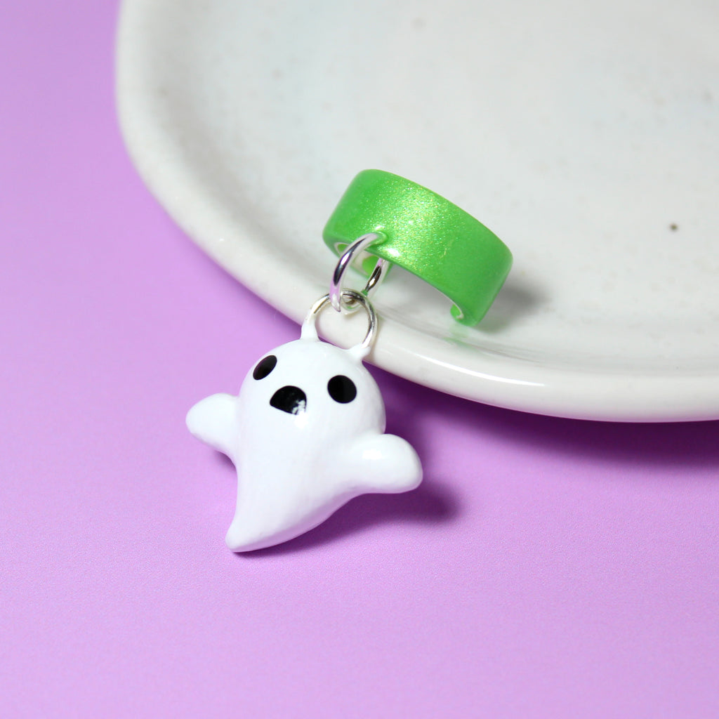 A ghost charm hangs from a sparkly green ear cuff. The ear cuff rests against a white jewelry dish.
