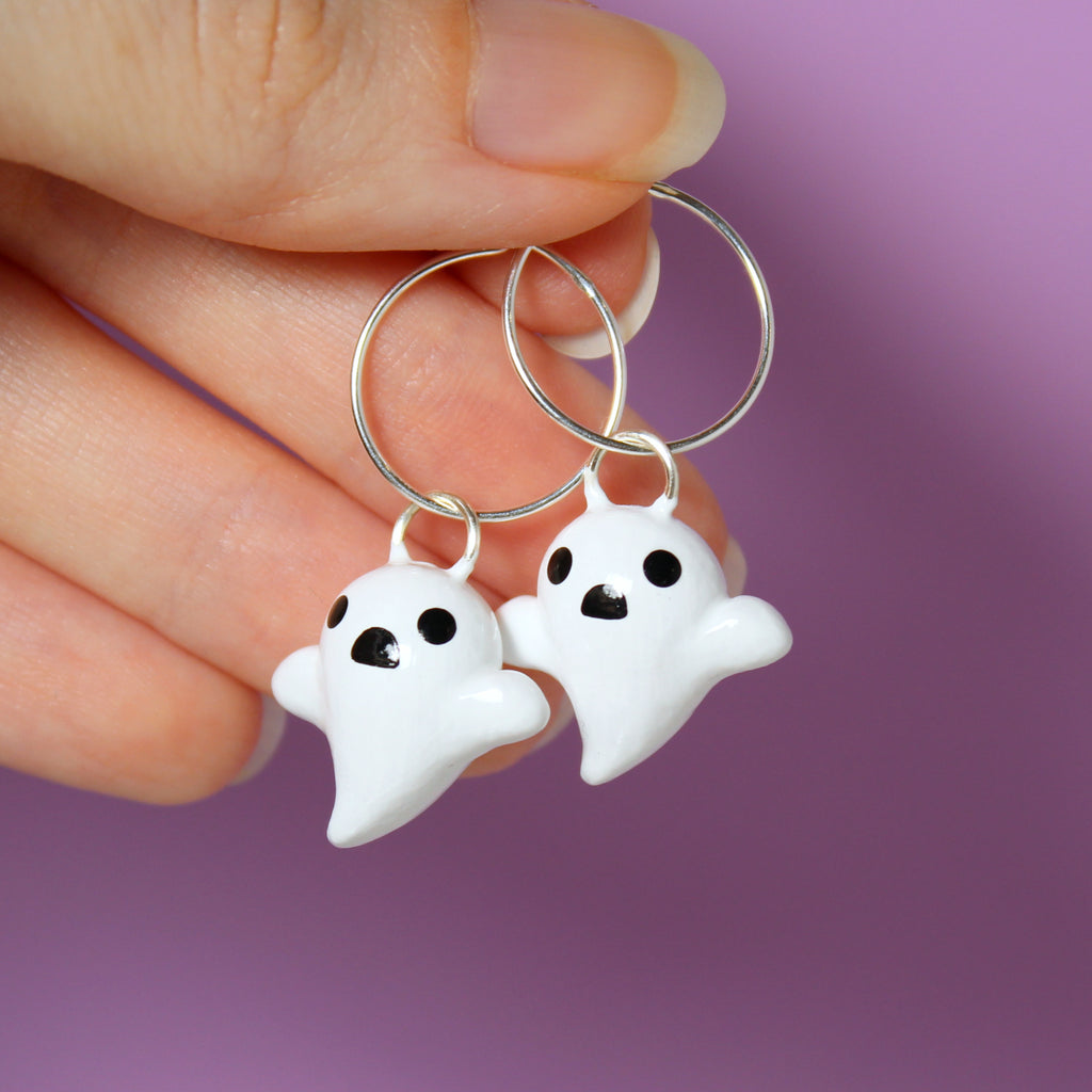 A hand holding a pair of ghost hoop earrings. The hoops are 19mm wide, and the ghost charms are about 3/4 of an inch tall.