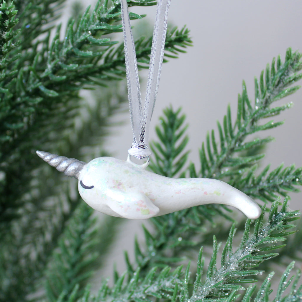 A white narwhal ornament with iridescent flakes hangs on a christmas tree.