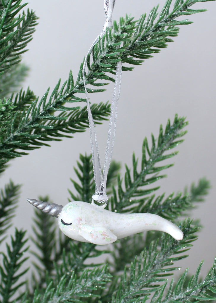 A white narwhal ornament with iridescent flakes hangs on a christmas tree.