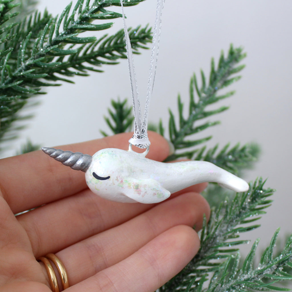 A hand holds a white narwhal ornament with shiny iridescent flakes. The narwhal is about 2.5 inches long.