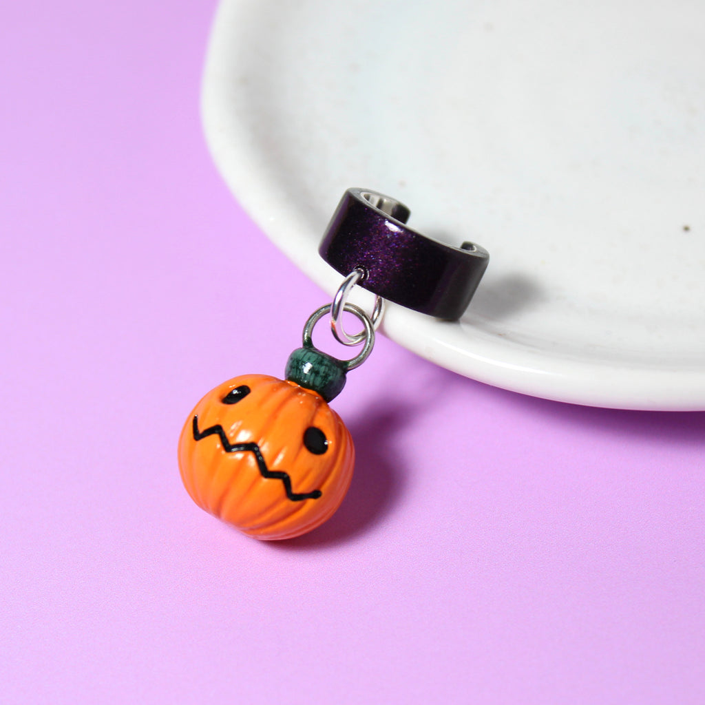 A jack o lantern charm hangs from a sparkly dark purple ear cuff. The ear cuff rests on a white jewelry dish.