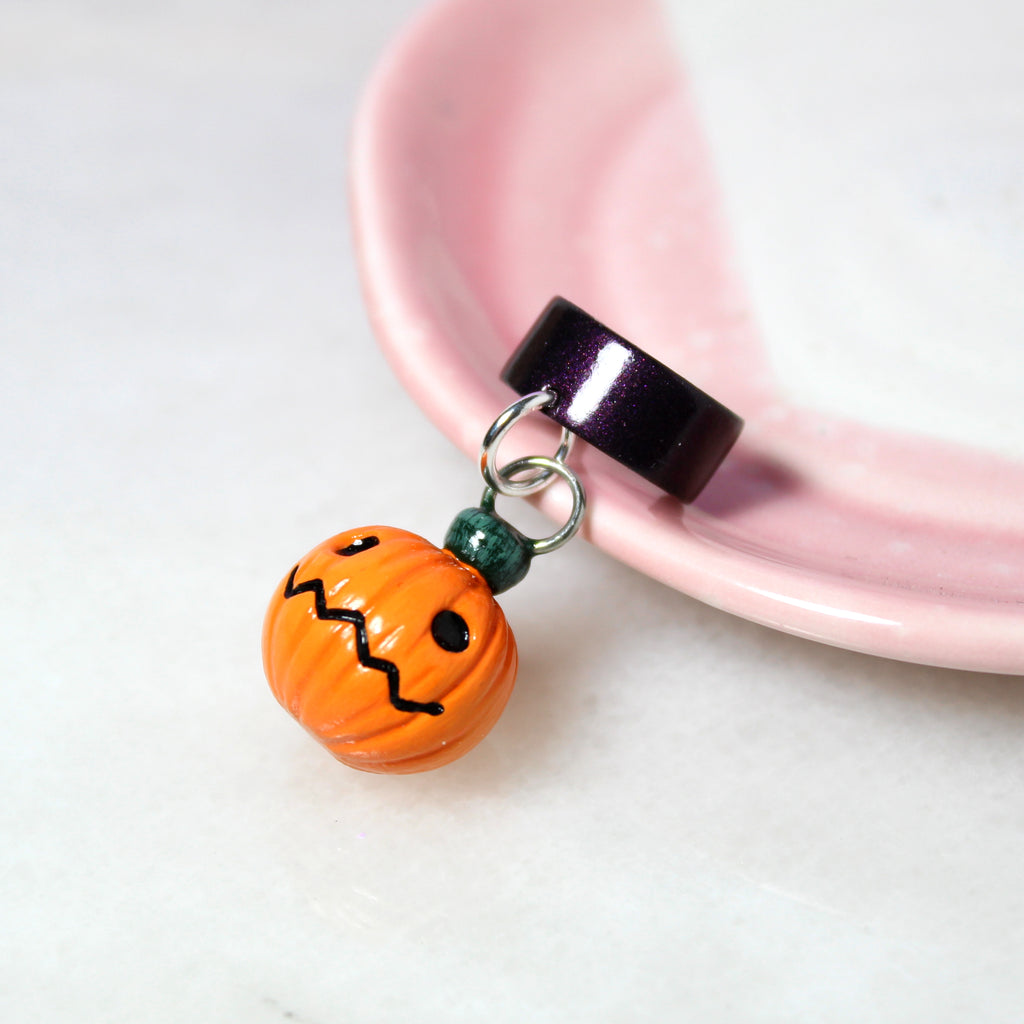 A jack o lantern charm hangs from a sparkly dark purple ear cuff. The ear cuff rests on a pink jewelry dish.
