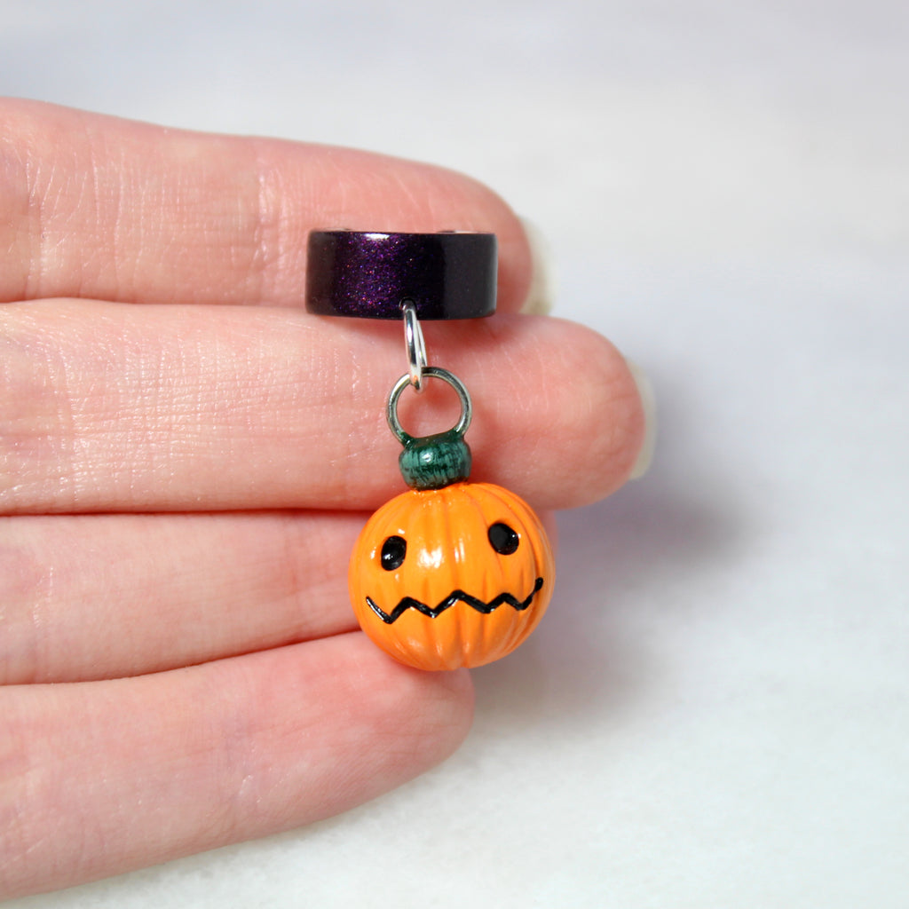 A hand holds a jack o lantern ear cuff. The jack o lantern charm is about 3/4 of an inch tall.