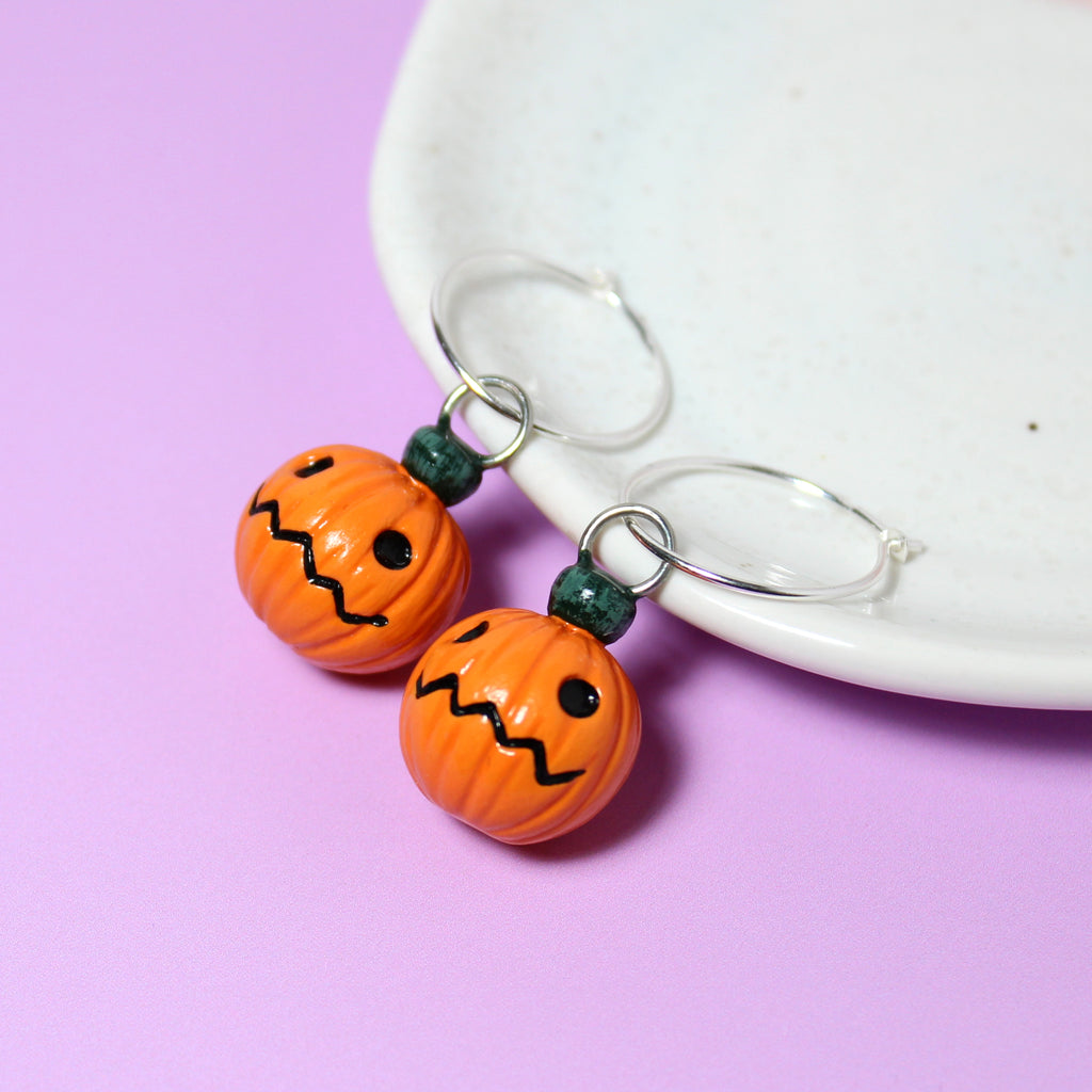 A pair of jack o lantern charms hanging from sterling silver hoop earrings. The earrings rest against a white jewelry dish.