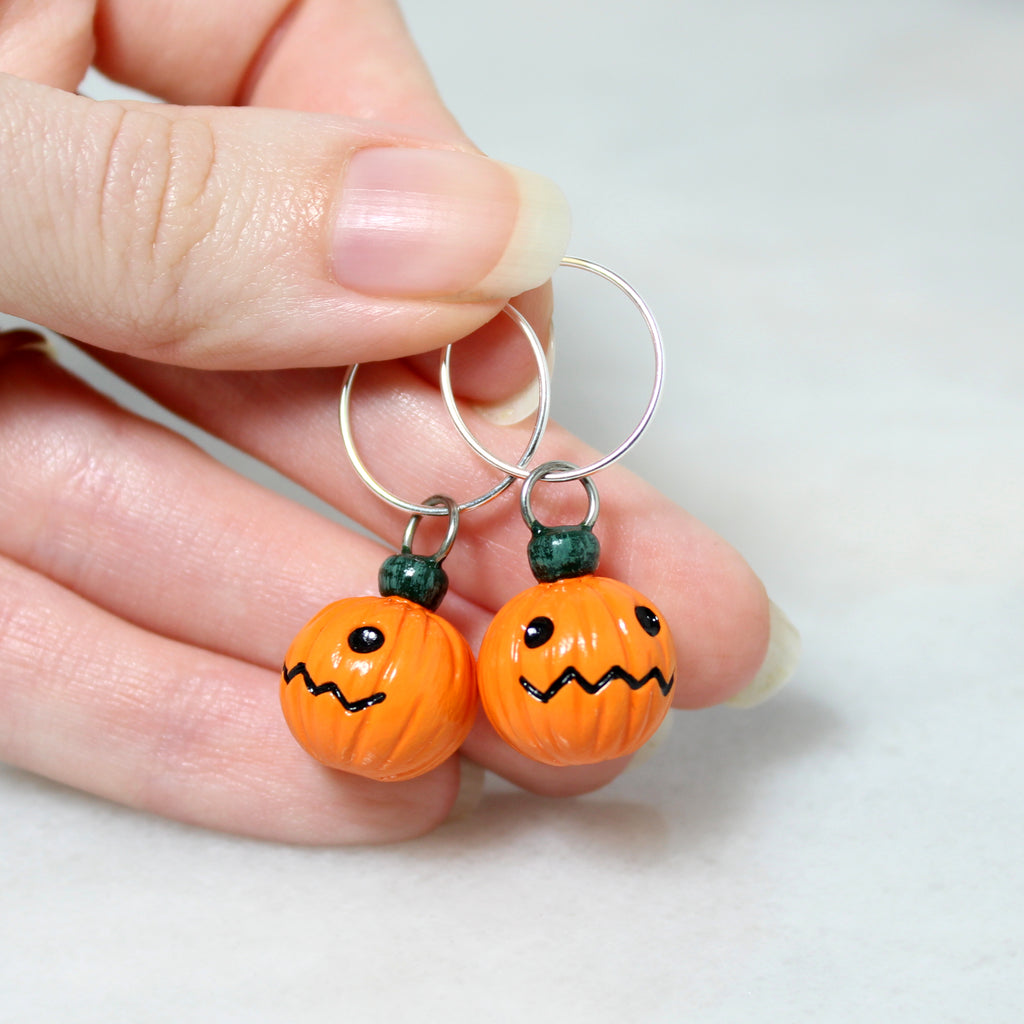 A hand holding a pair of jack o lantern hoop earrings. The sterling silver earrings are 19mm round, and the pumpkin charms are about 3/4 of an inch tall.