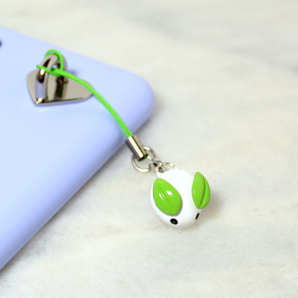 A white bunny charm with leafy green ears is attached to a green phone strap and hangs off of a phone case.