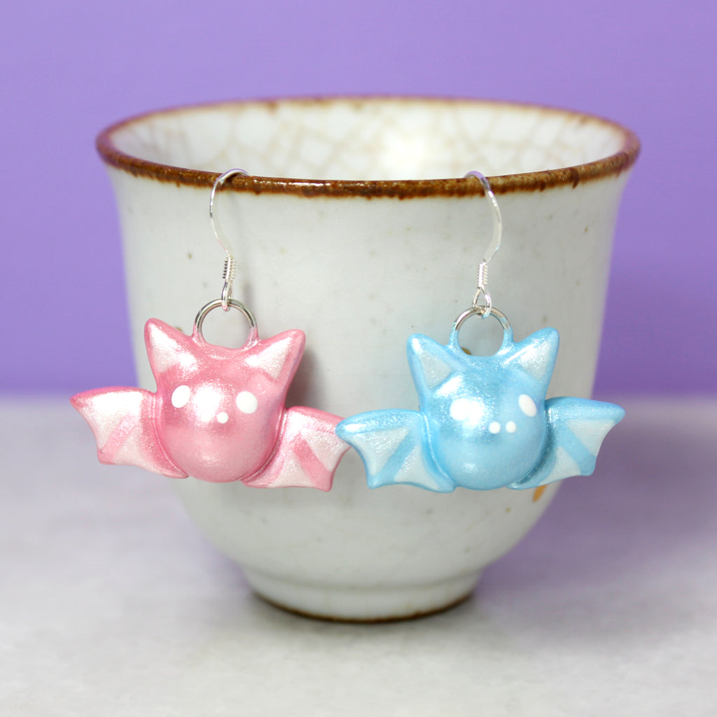A pair of pastel bat earrings hang from a white cup. The bat on the left is sparkly bubblegum pink, and the bat on the right is sparkly baby blue.