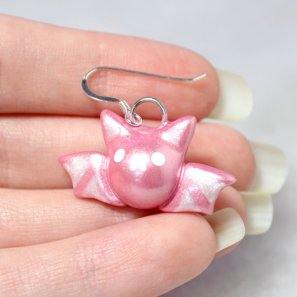 A hand holding a single sparkly pastel pink bat earring. The bat charm is about 3/4 of an inch tall and hangs from a sterling silver ear wire.