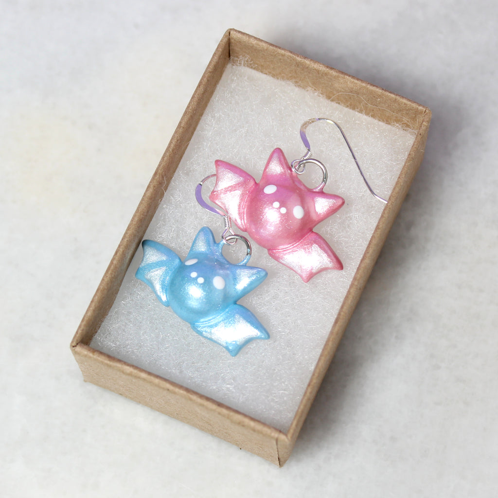 A pair of mismatched pastel bat earrings nestled in a kraft paper jewelry box.