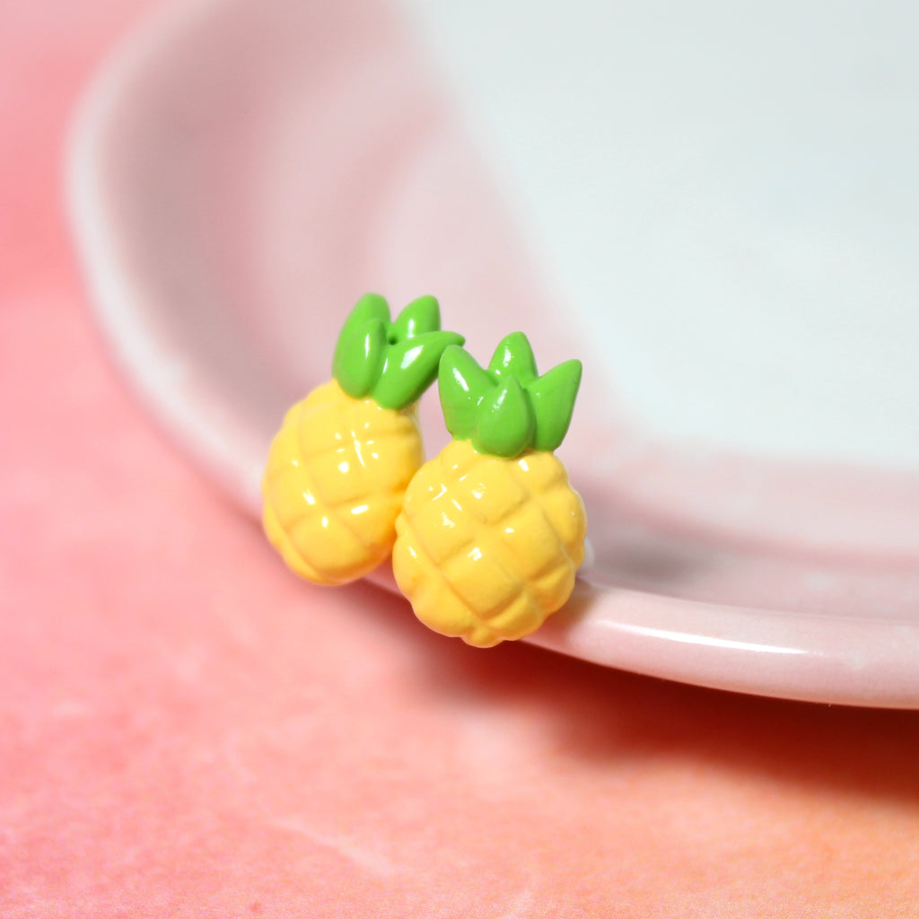 A pair of pineapple earrings rest on the rim of a pink and white dish. The dish sits on top of a watercolor pink background.