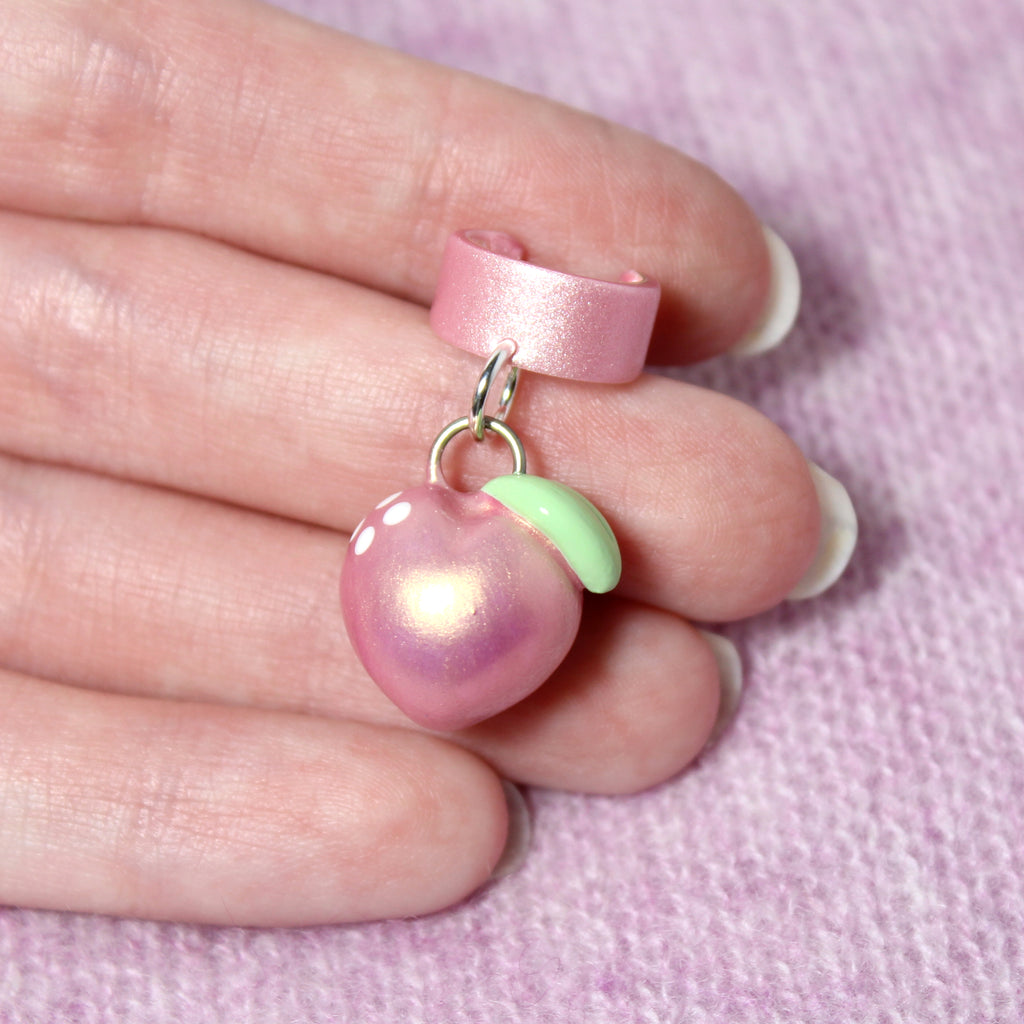A hand holds a pink gold peach ear cuff. The peach charm is about 3/4 of an inch tall.
