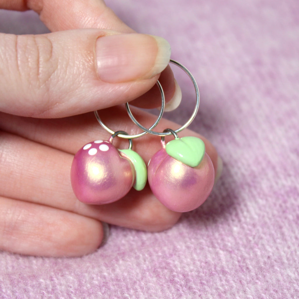 A hand holds a pair of pink gold peach hoop earrings. The hoops are about 19mm wide and the charms are 3/4 of an inch wide.