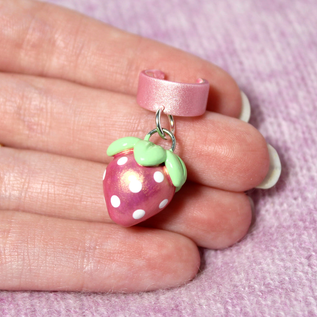 A hand holds a pink gold strawberry ear cuff. The strawberry charm is about 3/4 of an inch tall.