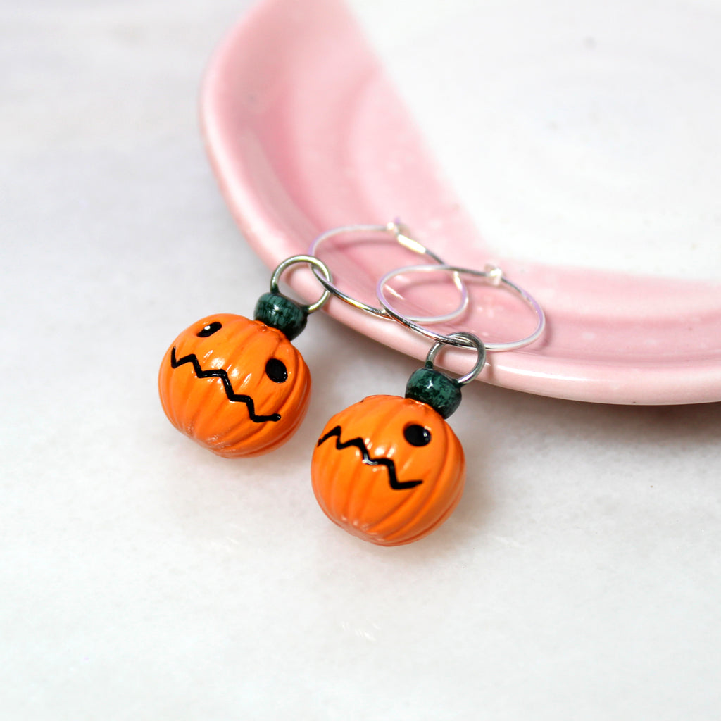 A pair of jack o lantern charms hanging from sterling silver hoop earrings. The earrings rest against a pink and white jewelry dish.